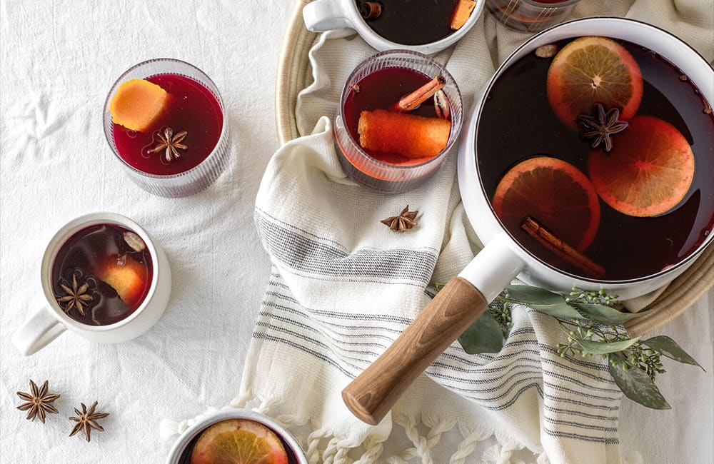 A cozy setup featuring cups of mulled wine with spices and citrus slices, alongside a pot on a tablecloth.