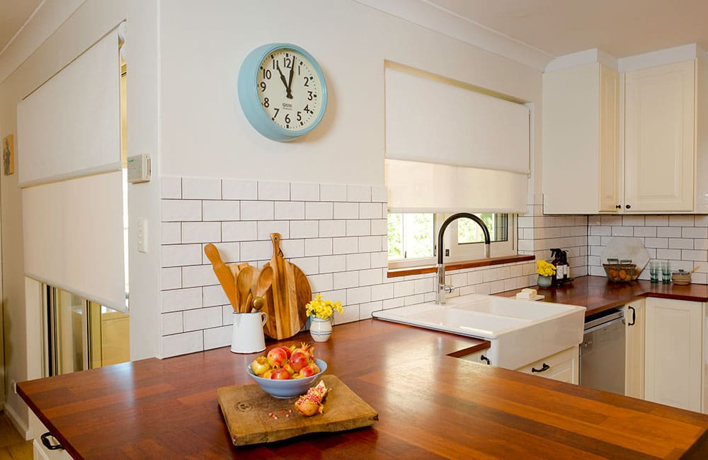 Bright modern kitchen with white blinds, wooden countertop, a bowl of apples, and a blue wall clock.