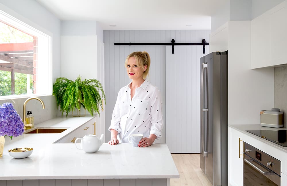 Bright kitchen with a woman in a white blouse standing by a countertop decorated with plants and a teapot.