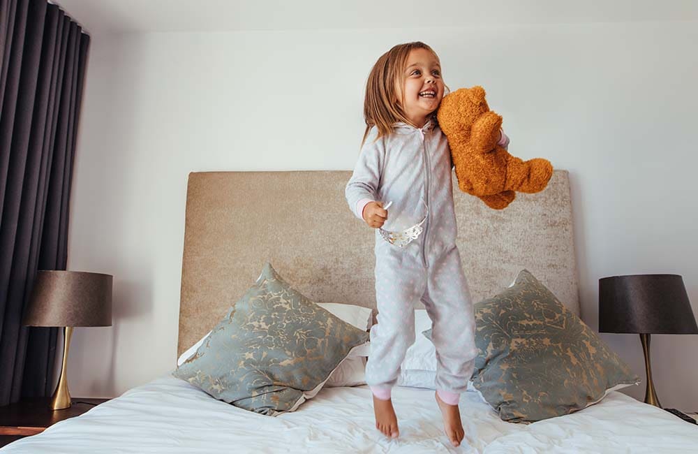 Happy toddler in pajamas jumping on a bed with a teddy bear, representing early morning energy.