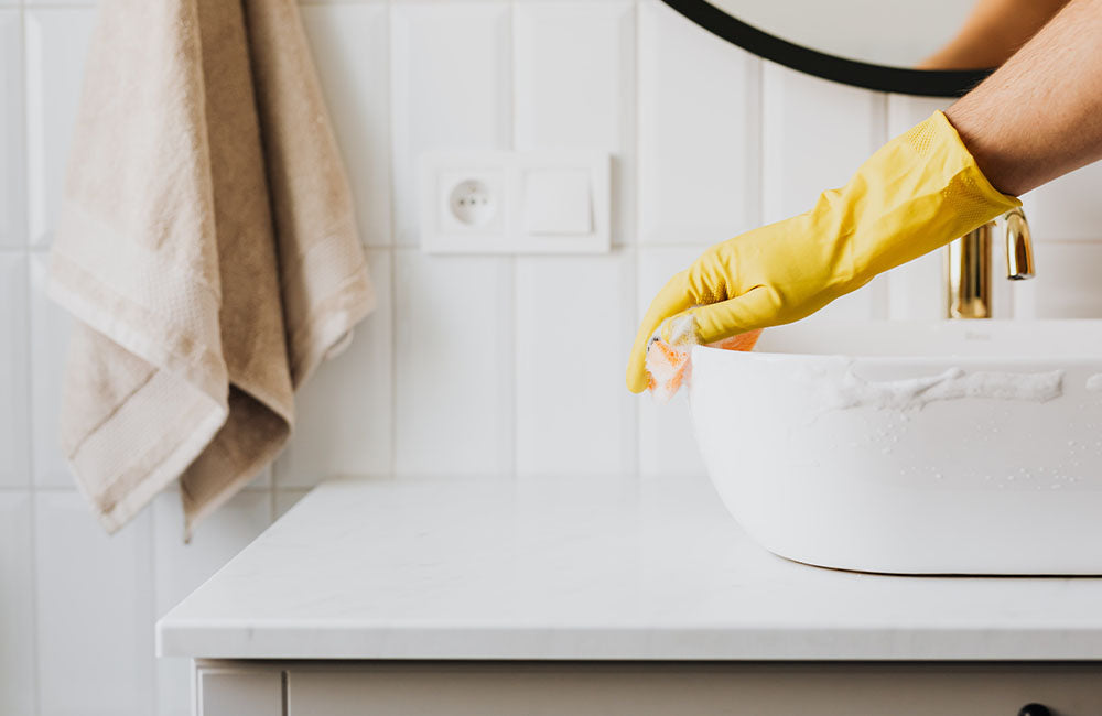 Person cleaning a white basin with a yellow rubber glove in a modern bathroom.