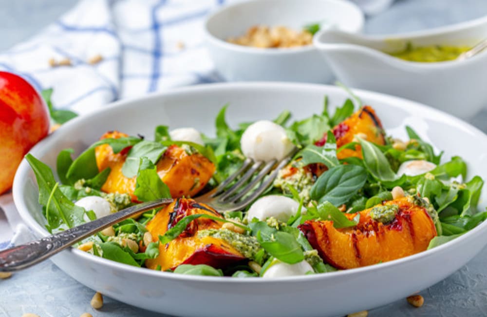 Grilled summer salad with peaches, mozzarella, arugula, and pine nuts served in a white bowl.