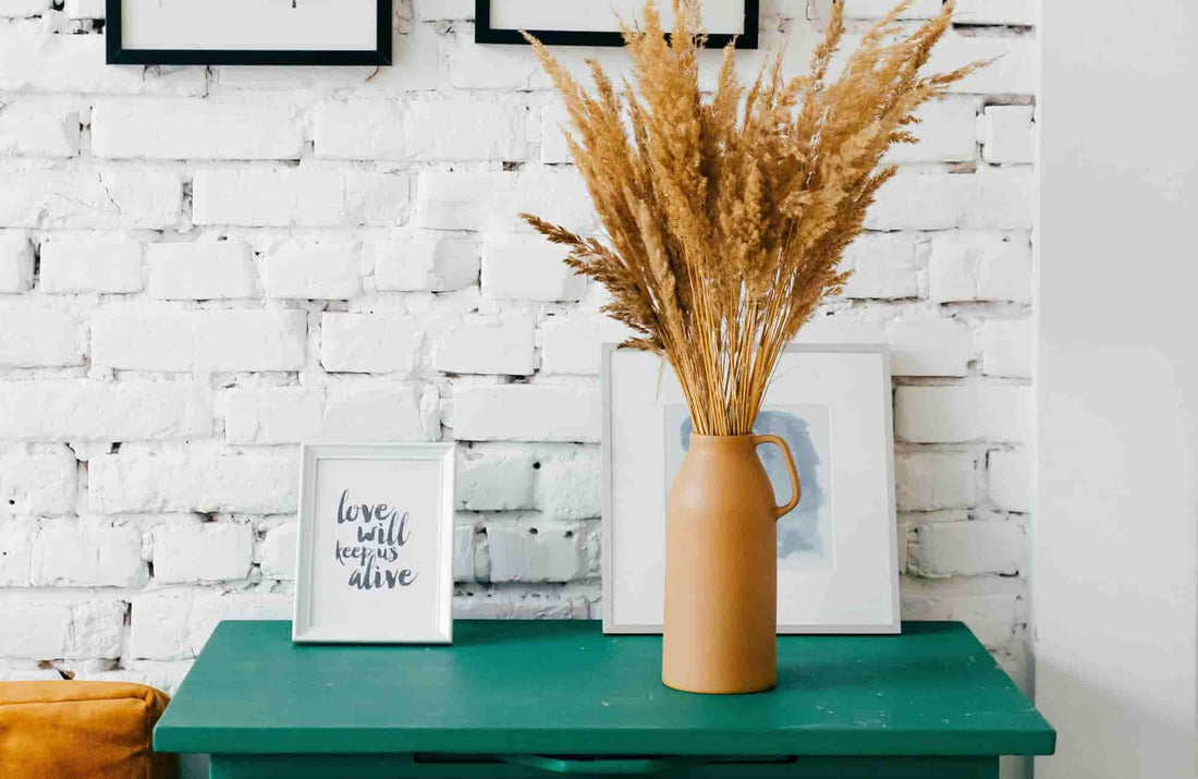 Stylish home decor featuring a vase with dried flowers on a green table, framed art, and a rustic white brick wall.